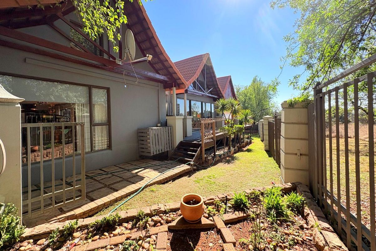 Rear exterior view featuring a paved patio, elevated timber deck, tiled roofline, and landscaped garden area enclosed by metal perimeter fencing.