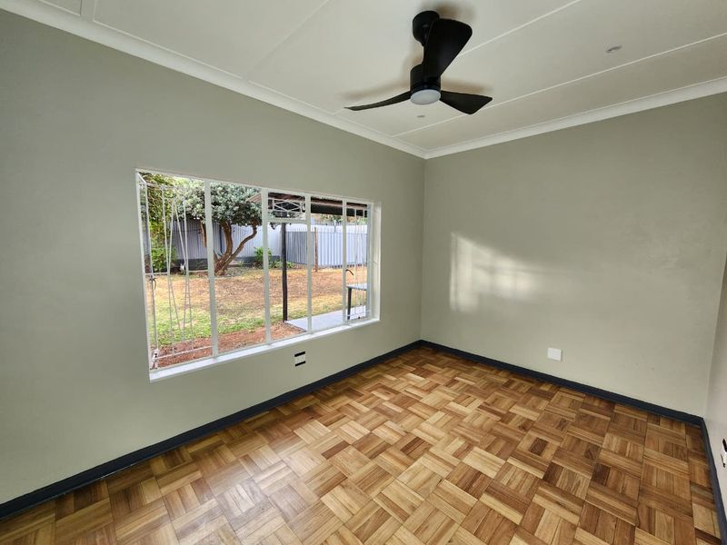 A bright, empty bedroom featuring well-maintained parquet flooring, a modern ceiling fan, and large windows with security bars overlooking the garden.