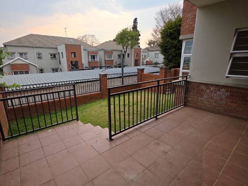 A covered patio area featuring tiled flooring, a metal safety balustrade, and brick wall detailing, overlooking a small private garden space.