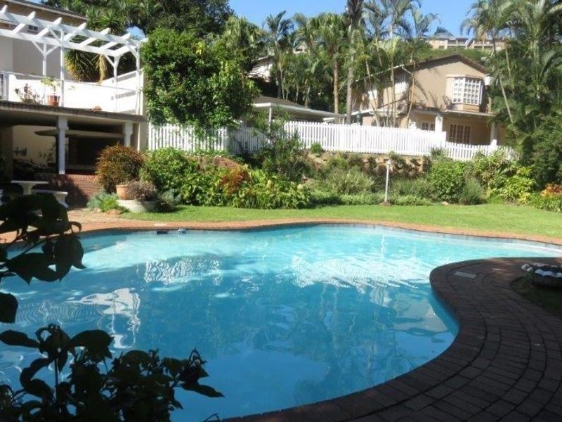 A well-maintained outdoor pool area featuring a swimming pool with brick coping, a pergola, and a landscaped garden with a white picket fence.