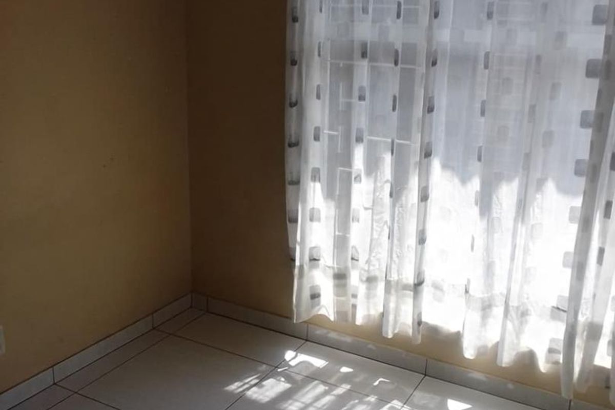 A bright, empty bedroom featuring tiled flooring and a large window with sheer curtains, offering a clean and neutral space.