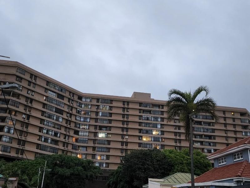 Street view showing the approach to a large multi-story apartment complex with surrounding residential properties and mature trees.