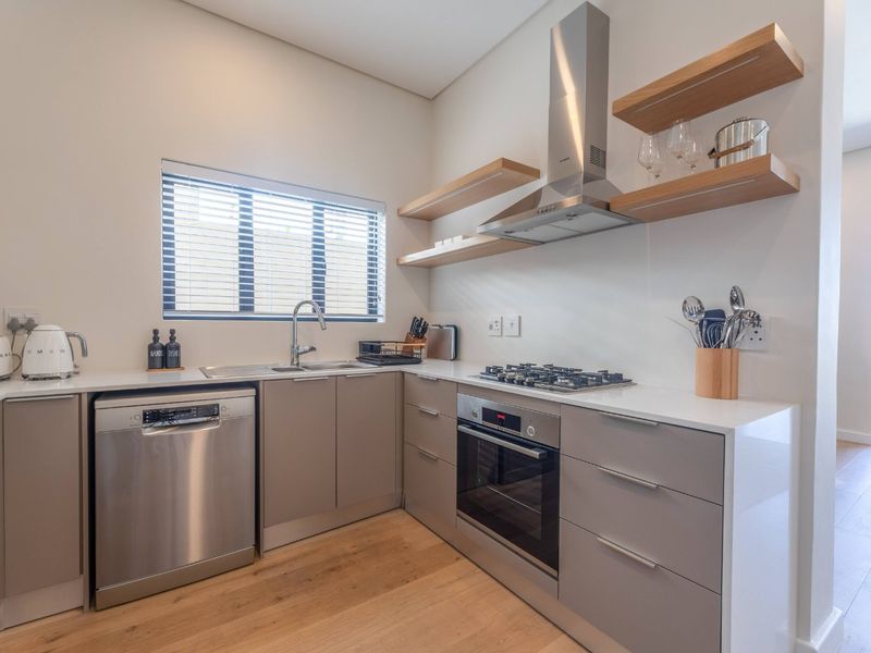 Modern kitchen featuring sleek grey cabinetry, stainless steel appliances, gas hob, built-in oven, and floating timber shelving with light laminate flooring.
