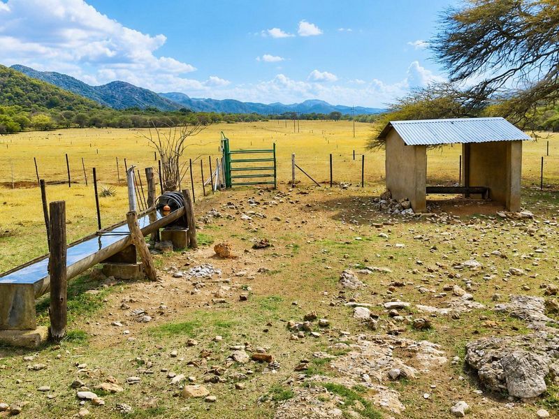Outdoor small holding area featuring a livestock water trough, a simple corrugated metal shelter, and wire fencing set against a scenic mountain landscape.
