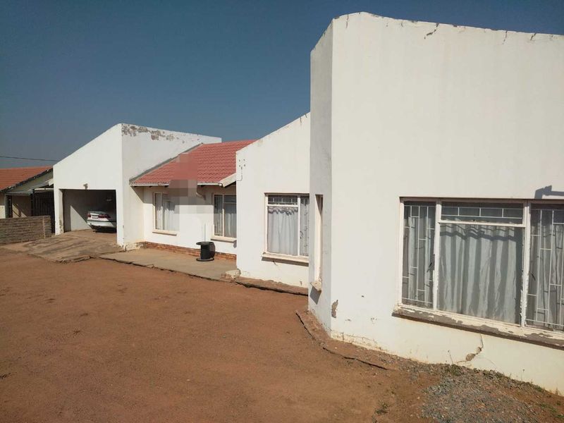 Front elevation of a single-story house featuring a tiled roof, metal-framed windows, and an attached carport, showing signs of exterior wear and plaster cracking.