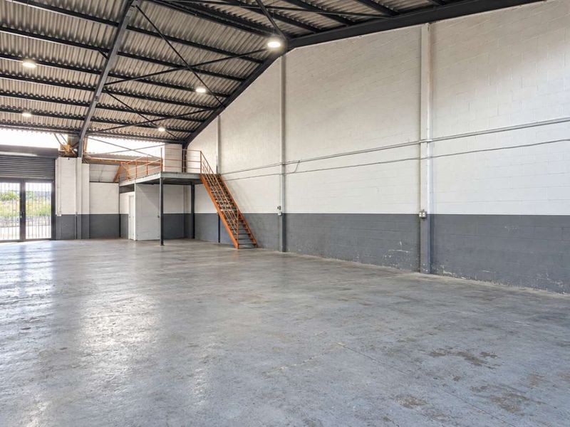 Spacious industrial warehouse unit featuring high ceilings with exposed steel trusses, concrete flooring, a mezzanine level with staircase, and a roller shutter door.