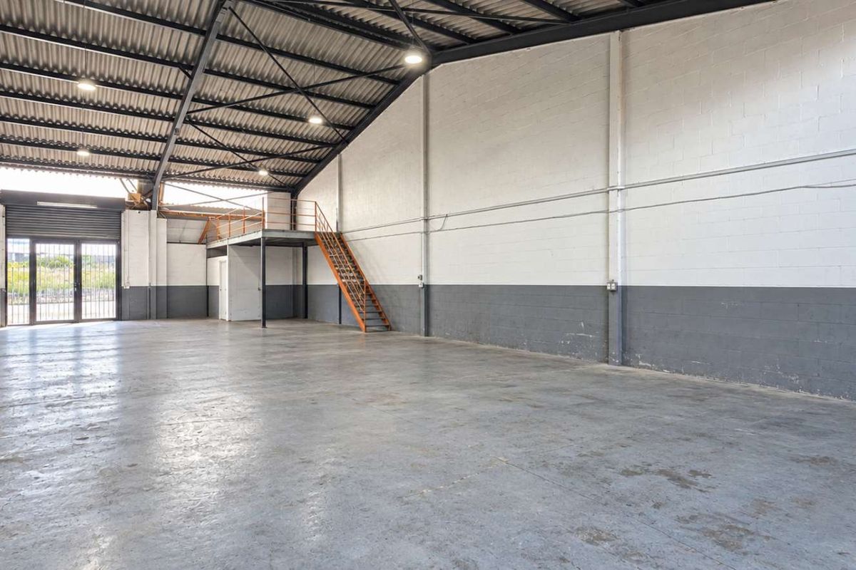 Spacious industrial warehouse unit featuring high ceilings with exposed steel trusses, concrete flooring, a mezzanine level with staircase, and a roller shutter door.