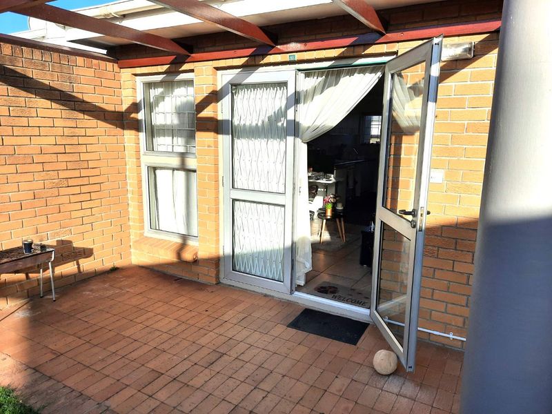 A paved outdoor patio area featuring a timber pergola, brick facade, and aluminium-framed glass entrance door.