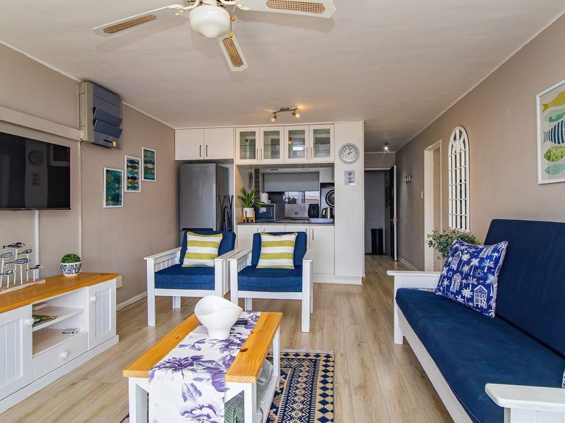 Open-plan living area featuring laminate flooring, a ceiling fan, wall-mounted air conditioning, and a compact integrated kitchen with built-in cabinetry.