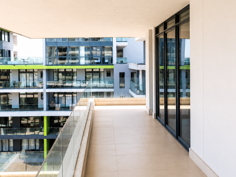 A spacious, modern covered balcony featuring tiled flooring, a sleek glass balustrade, and large sliding glass doors providing access to the apartment interior.