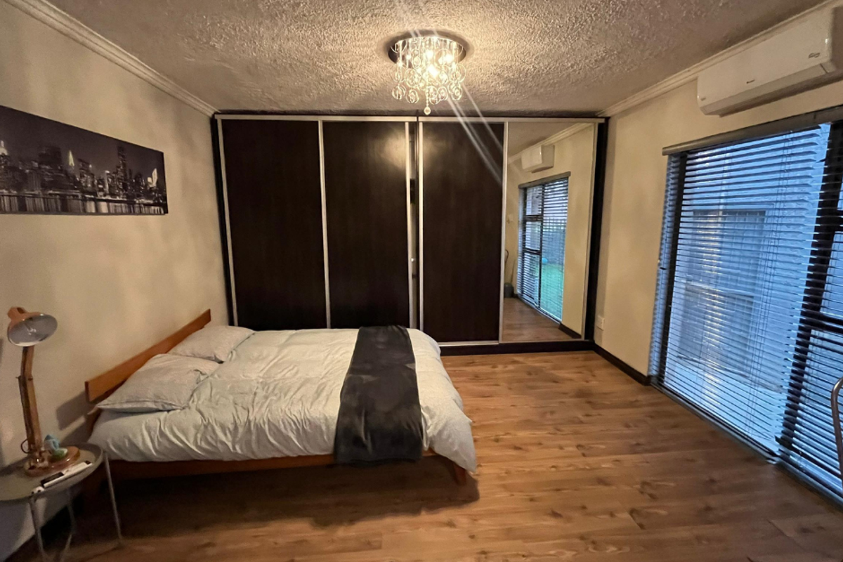 Spacious bedroom featuring laminate flooring, built-in mirrored wardrobes, and a wall-mounted split-system air conditioner with large windows and venetian blinds.