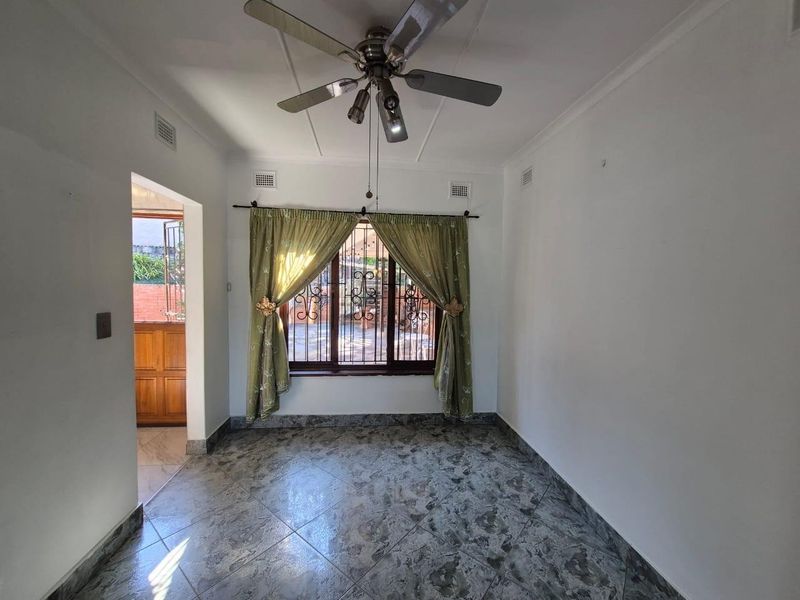An empty living room featuring tiled flooring, a ceiling fan, and security bars on the window, with an adjacent doorway leading to an outdoor area.