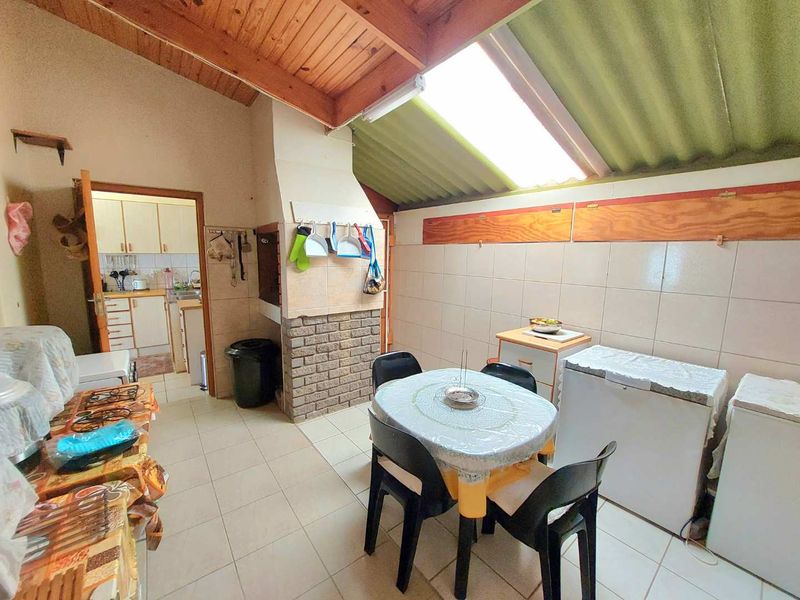 A dated kitchen and dining area featuring a built-in braai, tiled flooring, and an exposed timber ceiling with a skylight.