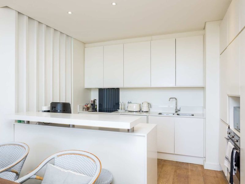 Modern, minimalist kitchen featuring sleek white cabinetry, stone countertops, a breakfast bar, integrated oven, and high-quality timber flooring.