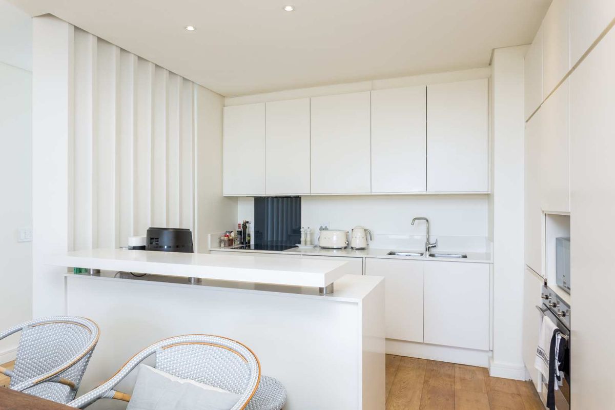 Modern, minimalist kitchen featuring sleek white cabinetry, stone countertops, a breakfast bar, integrated oven, and high-quality timber flooring.