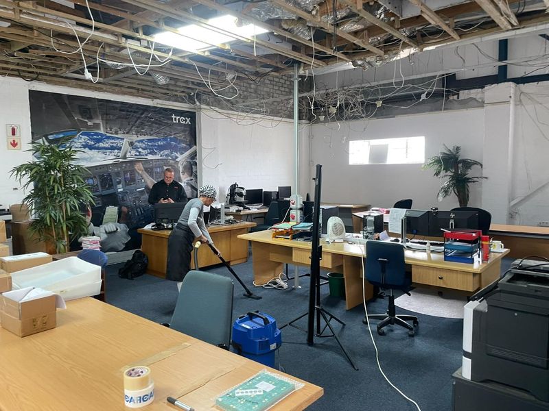 Open-plan office space featuring exposed ceiling structure, exposed electrical wiring, and carpeted flooring, currently undergoing maintenance.