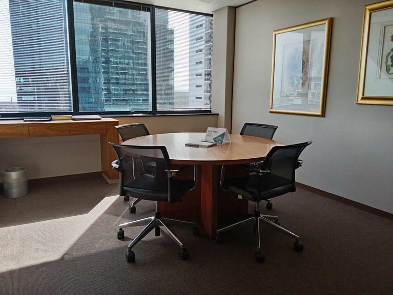 A professional office space featuring large floor-to-ceiling windows with city views, built-in wooden desk, and carpeted flooring.