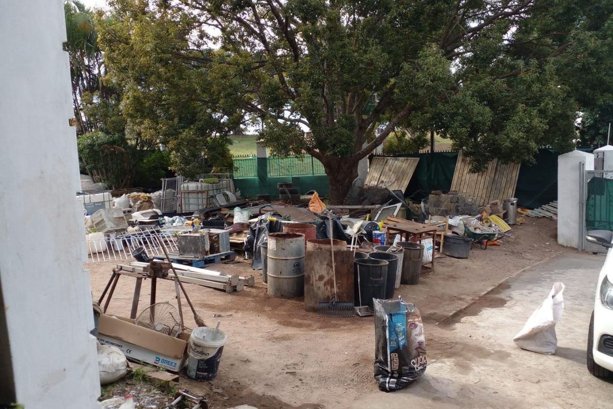 An outdoor rear area featuring a large mature tree and perimeter fencing, currently cluttered with significant amounts of debris and waste materials.