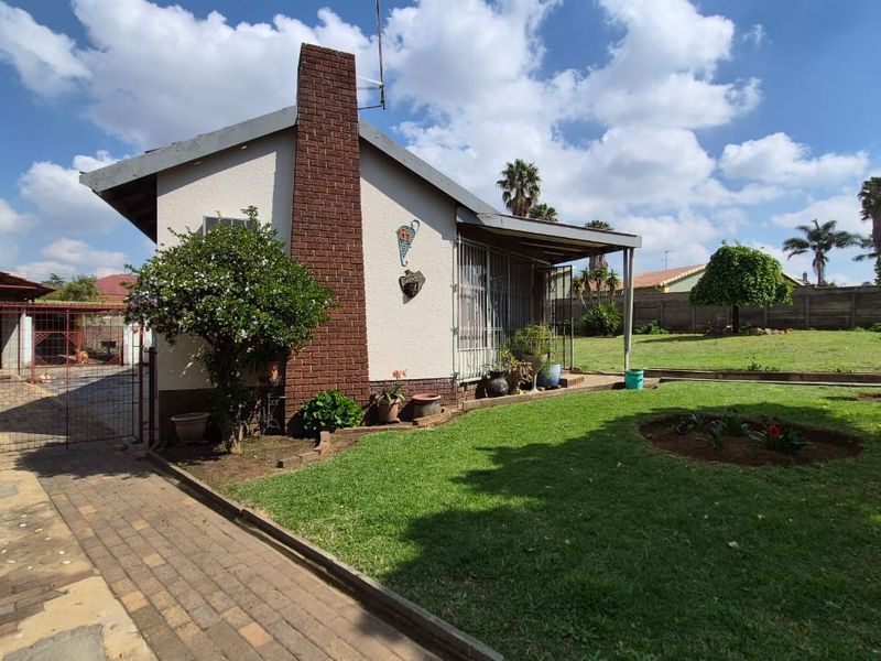 Rear view of the property featuring a brick chimney, covered patio area, paved driveway, and a spacious garden with perimeter walling.