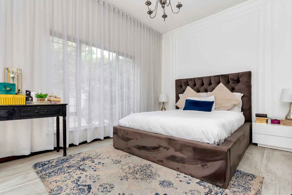 A well-maintained bedroom featuring elegant wall molding, large windows with sheer curtains, and a modern chandelier, creating a bright and sophisticated living space.