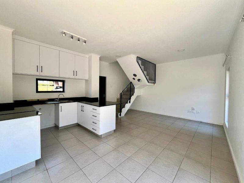 An open-plan kitchen and living area featuring modern white cabinetry, black granite countertops, tiled floors, and a staircase with a sleek metal balustrade.