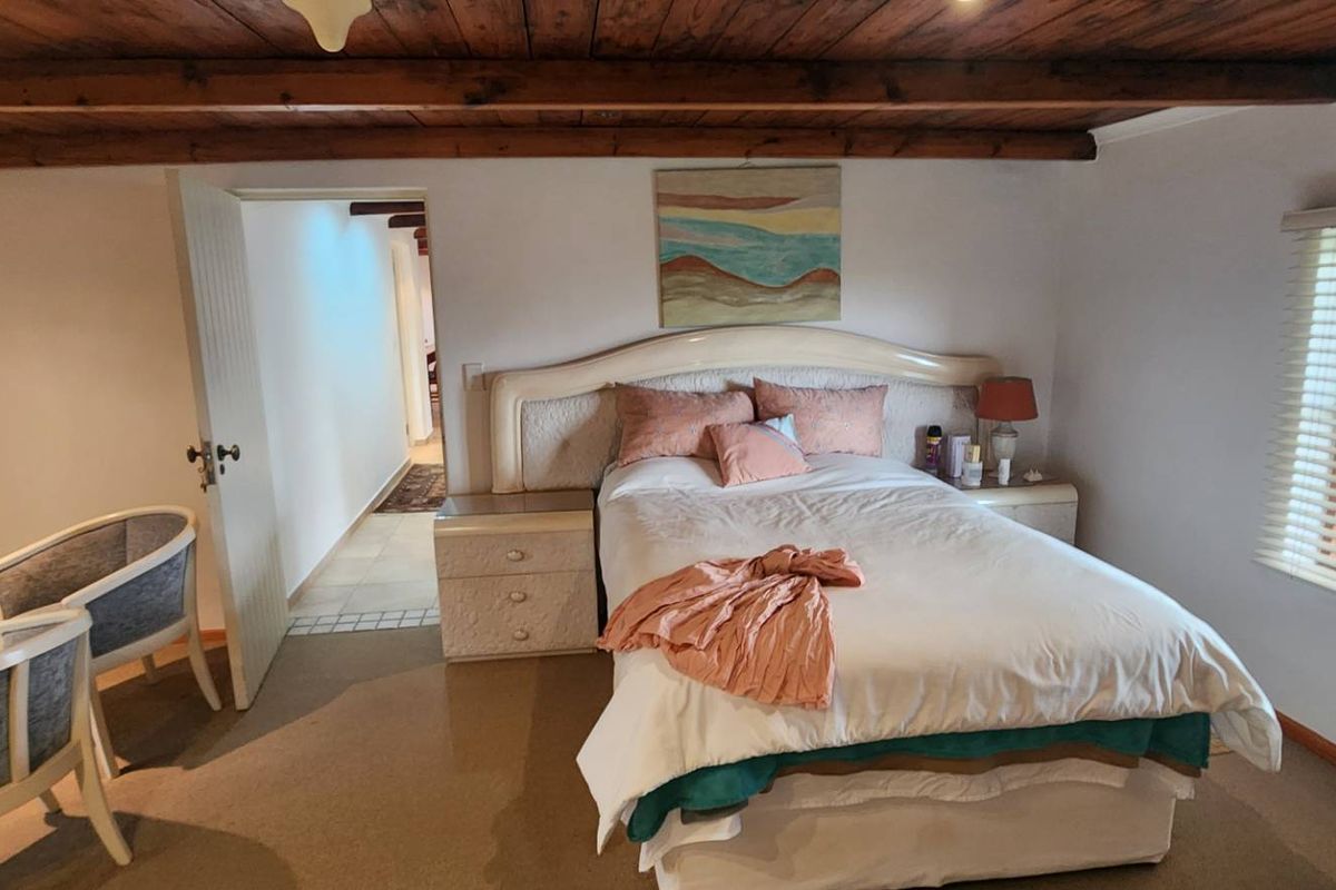 A cozy bedroom featuring exposed timber ceiling beams, carpeted flooring, and natural light through venetian blinds, leading into an adjacent hallway.