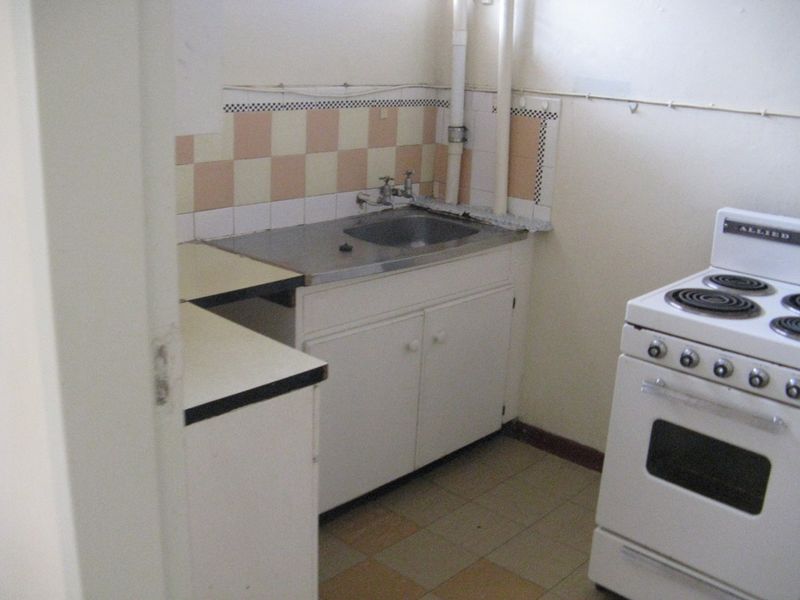 A dated kitchen featuring original cabinetry, a stainless steel sink, tiled splashback, and exposed plumbing, offering significant potential for a modern renovation.