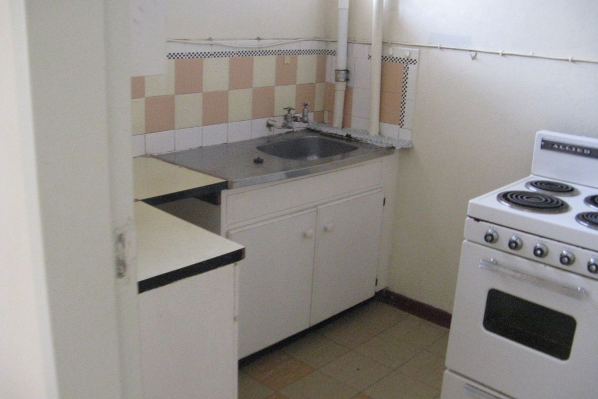 A dated kitchen featuring original cabinetry, a stainless steel sink, tiled splashback, and exposed plumbing, offering significant potential for a modern renovation.