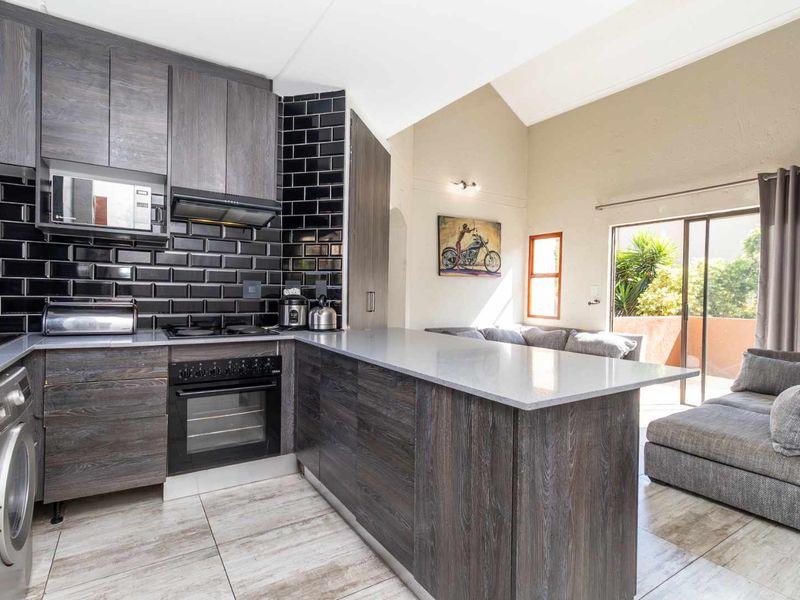 Modern open-plan kitchen featuring dark wood cabinetry, stone countertops, and a subway tile backsplash, flowing into a bright living area with vaulted ceilings.
