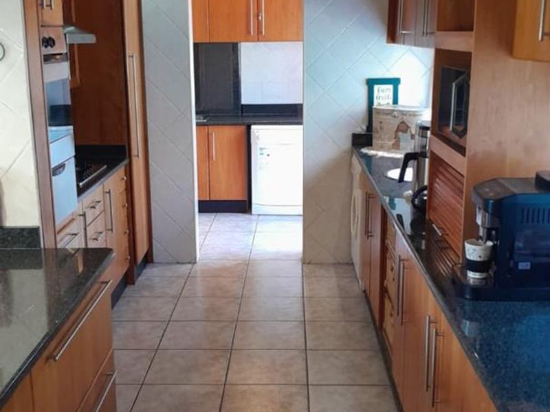 A galley-style kitchen featuring warm-toned wooden cabinetry, granite countertops, built-in oven, and tiled flooring, with an adjacent scullery area visible through the doorway.