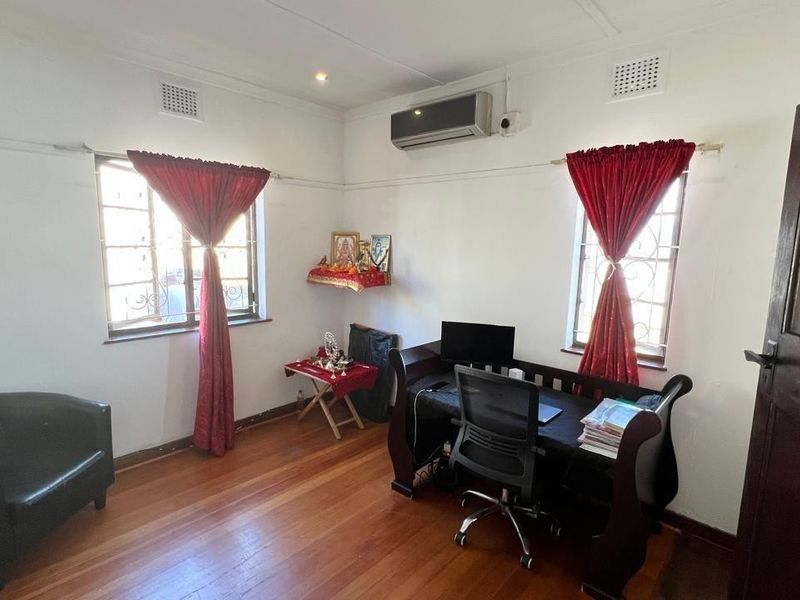 A functional home office featuring polished hardwood flooring, dual windows with security bars, and a wall-mounted split-system air conditioning unit.