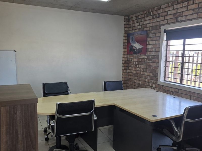 A functional office space featuring an exposed concrete ceiling, brick accent wall, tiled flooring, and large window with security bars.