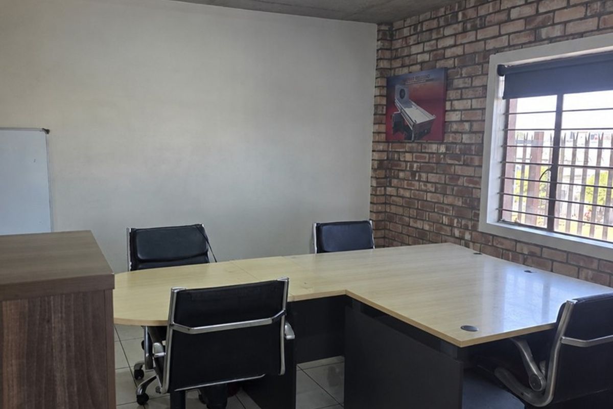 A functional office space featuring an exposed concrete ceiling, brick accent wall, tiled flooring, and large window with security bars.