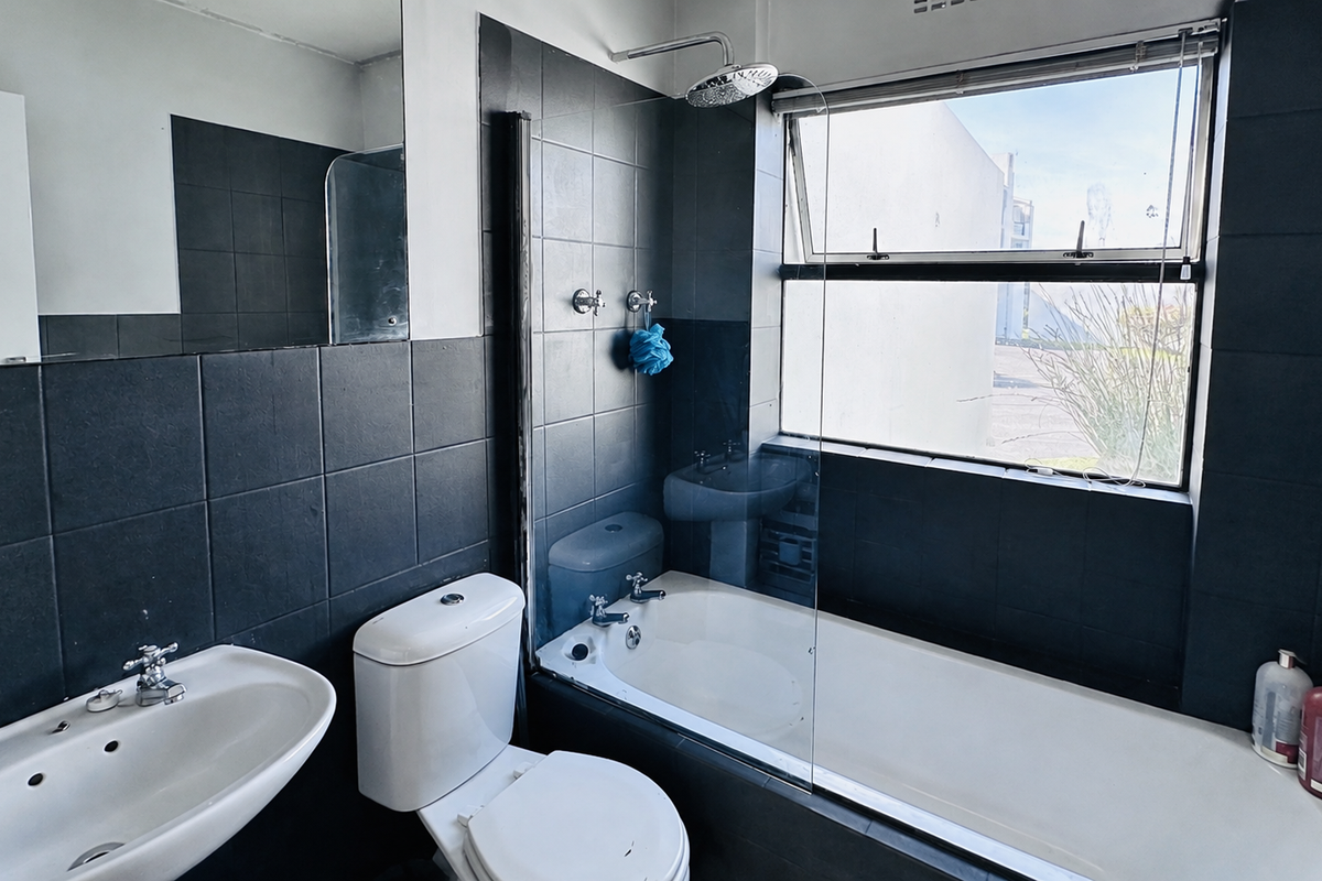 A modern bathroom featuring dark tiled walls, a white bathtub with a glass screen, a wall-mounted basin, and a toilet.