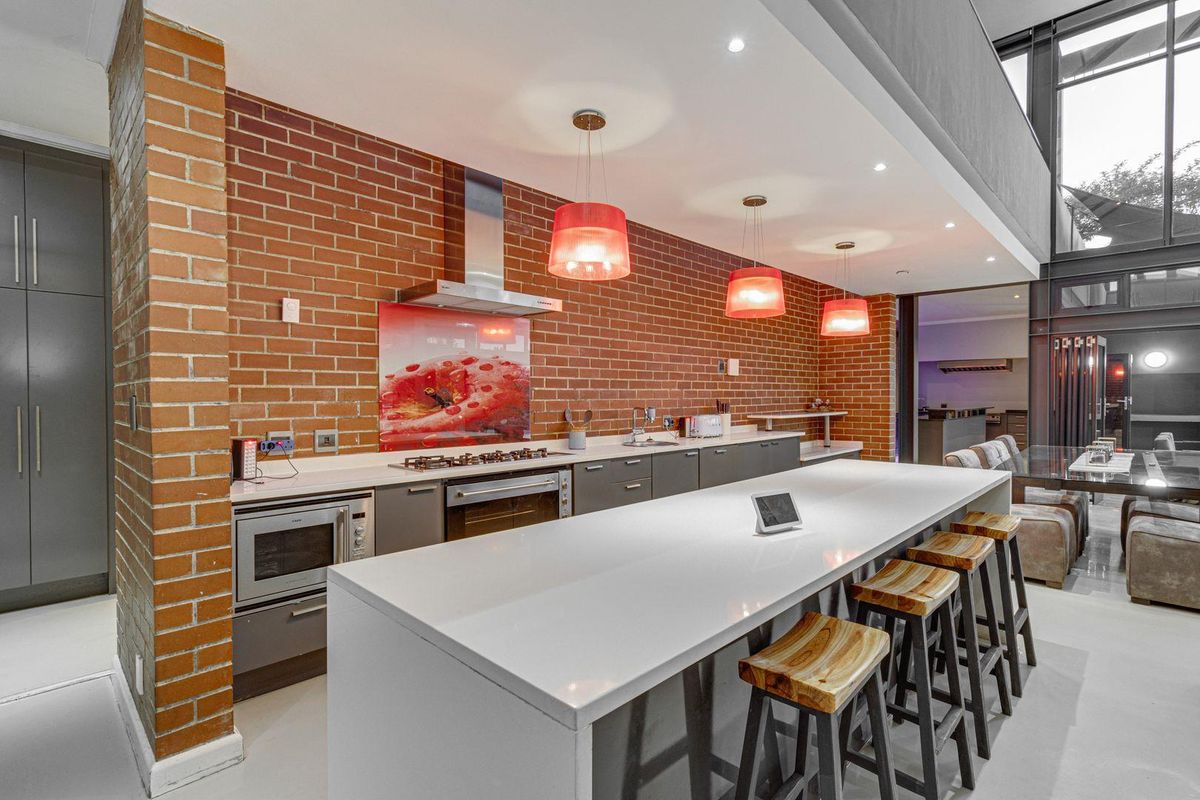 Modern open-plan kitchen featuring a large central island with stone countertop, exposed brick feature wall, built-in oven, gas hob, and stainless steel extractor fan.