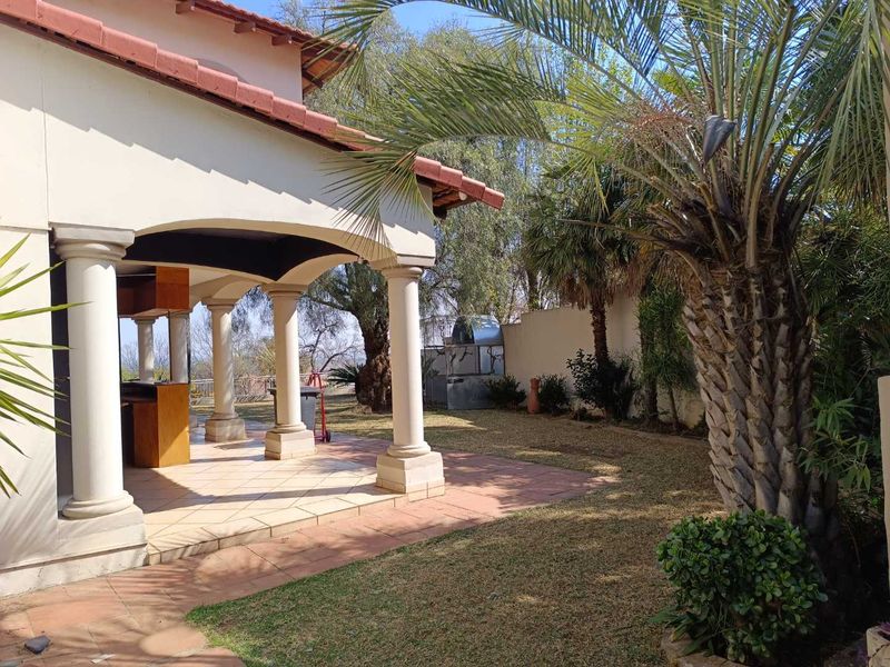 A covered outdoor patio area featuring classic columns, tiled flooring, and a built-in bar, overlooking a landscaped garden with mature palm trees.
