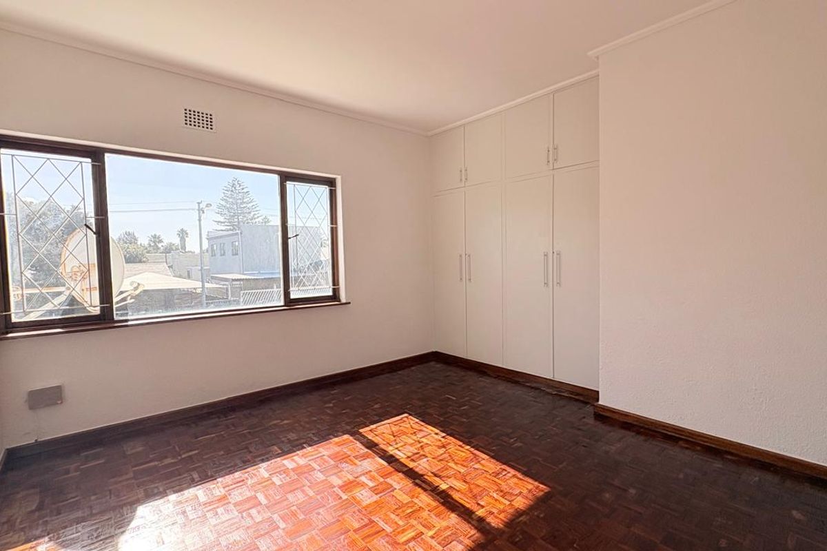 A bright, empty bedroom featuring original parquet flooring, built-in floor-to-ceiling wardrobes, and large windows with security bars.