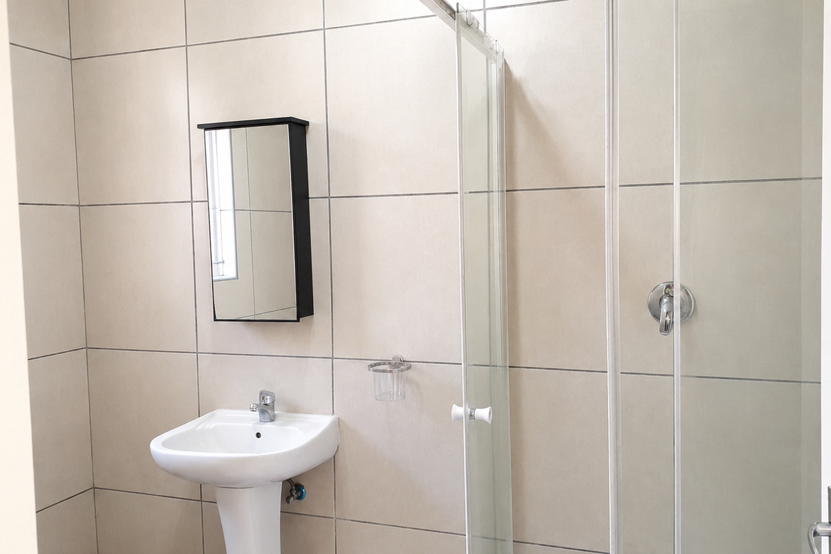 A clean, modern bathroom featuring a pedestal basin, glass shower enclosure, and neutral wall tiling.