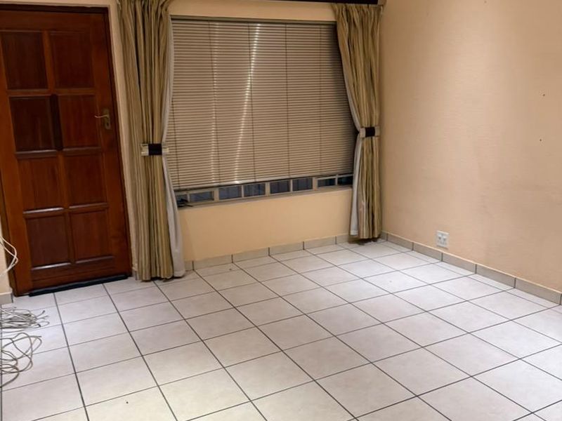 An empty living room featuring neutral tiled flooring, a ceiling fan, and a timber panel door with natural light provided by a window with blinds.