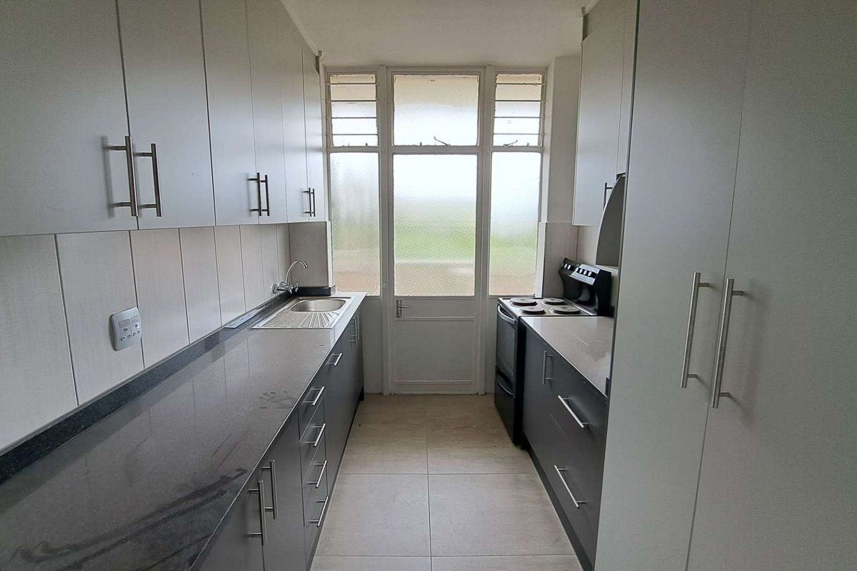 A galley-style kitchen featuring dark cabinetry, stone-look countertops, a stainless steel sink, and a freestanding electric stove with an integrated extractor fan.