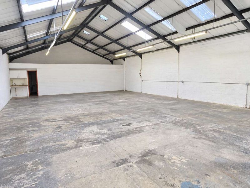 Spacious open-plan office or industrial unit featuring exposed steel roof trusses, skylights, painted brick walls, and a small kitchenette area.