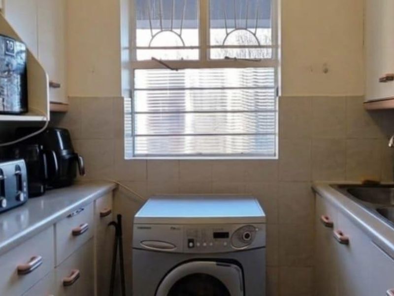 A compact kitchen featuring built-in cabinetry, a double stainless steel sink, tiled backsplash, and a window with security bars.