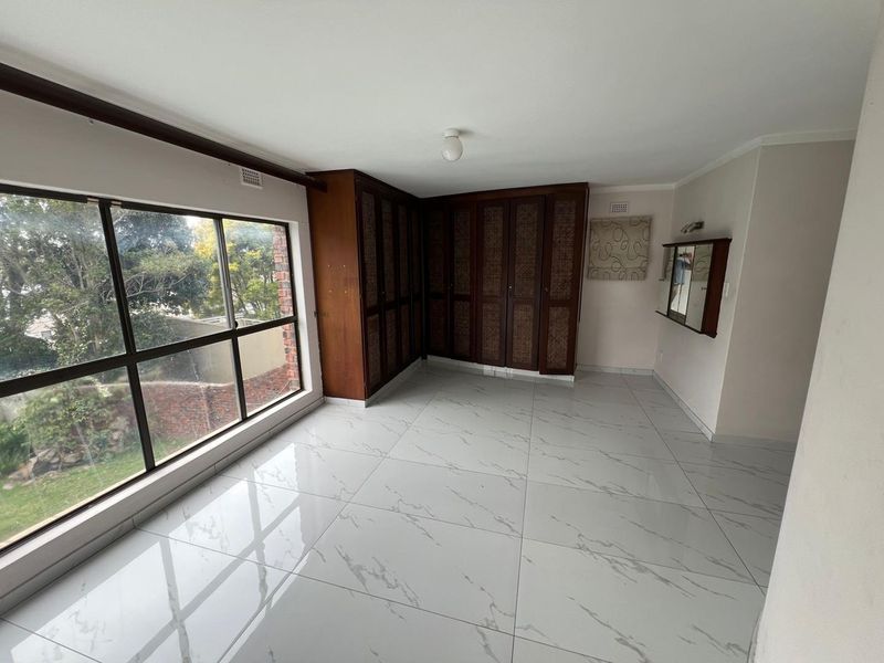 A bright, empty bedroom featuring modern white marble-look floor tiles, large windows providing natural light, and extensive built-in wooden cabinetry.