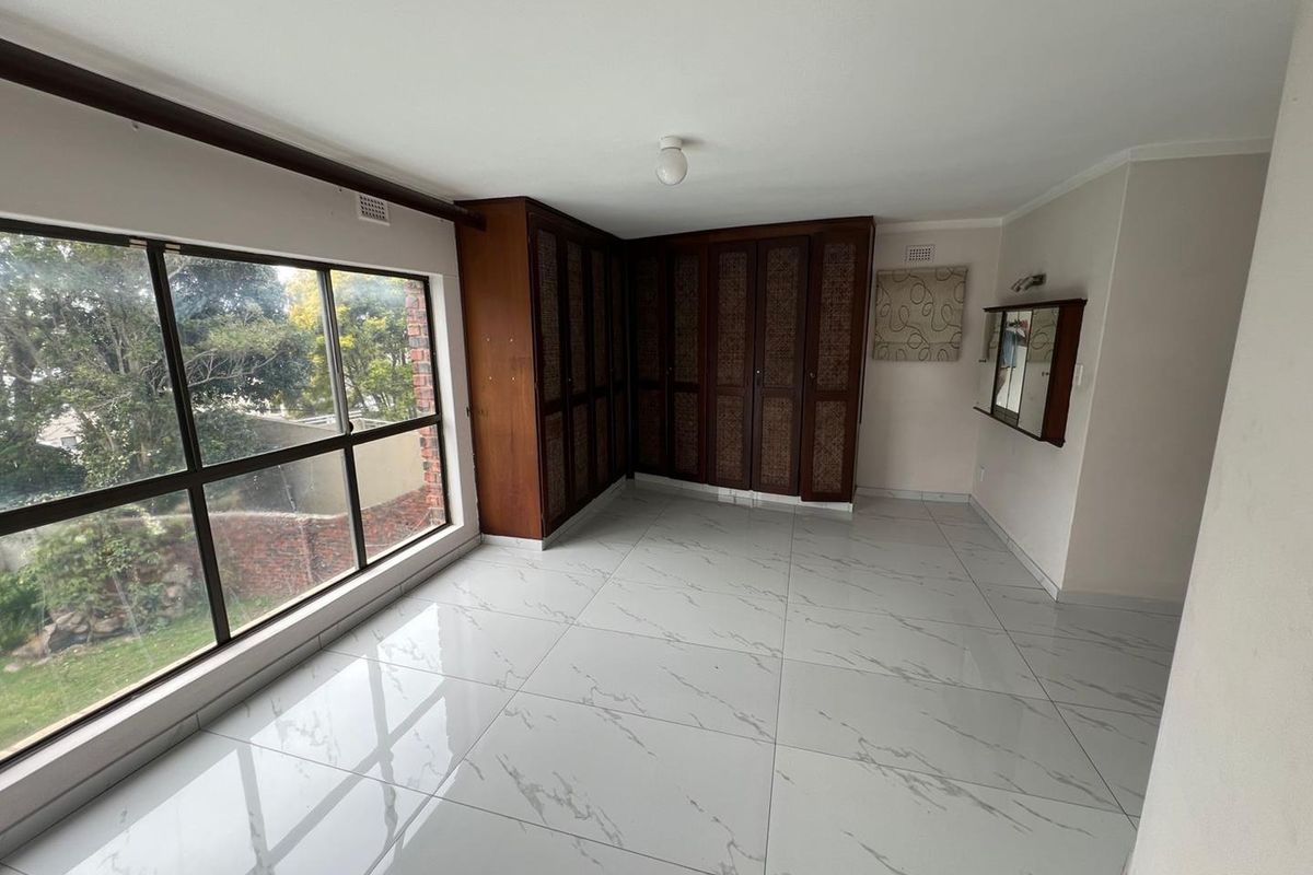 A bright, empty bedroom featuring modern white marble-look floor tiles, large windows providing natural light, and extensive built-in wooden cabinetry.