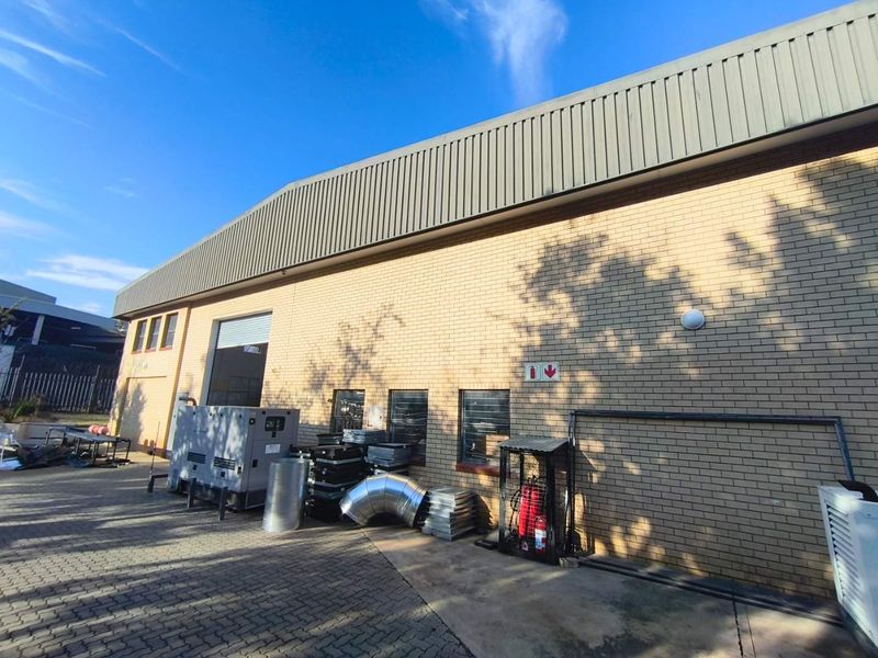Rear exterior view of an industrial warehouse building featuring brick masonry, metal cladding, a roller shutter door, and a paved yard area.