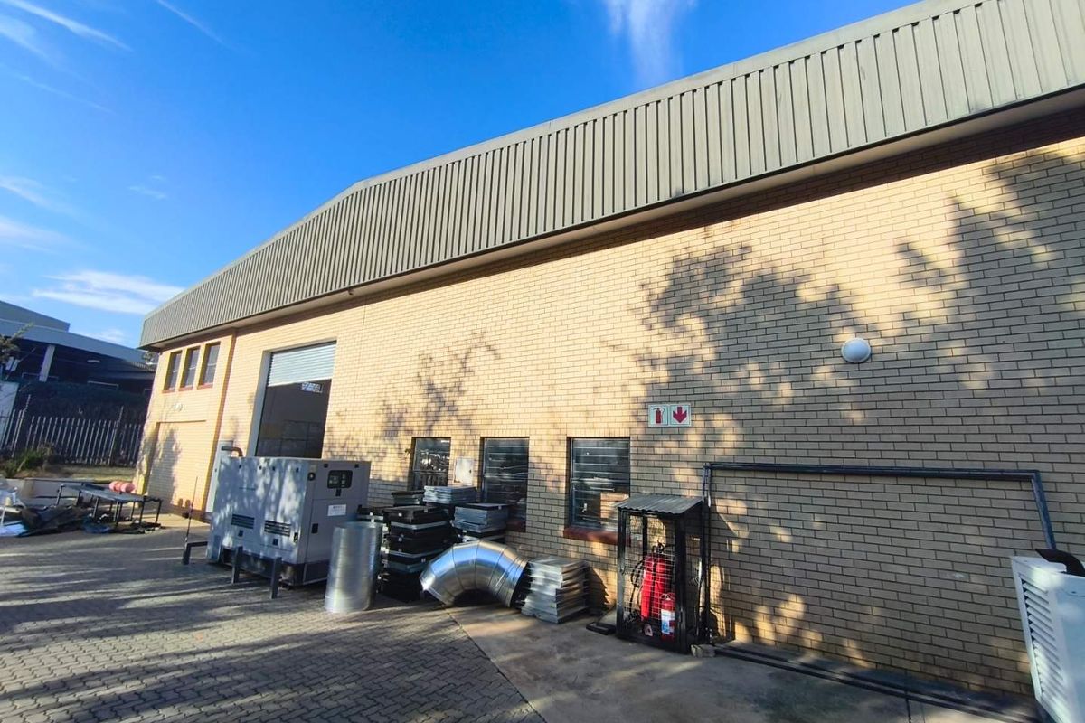 Rear exterior view of an industrial warehouse building featuring brick masonry, metal cladding, a roller shutter door, and a paved yard area.