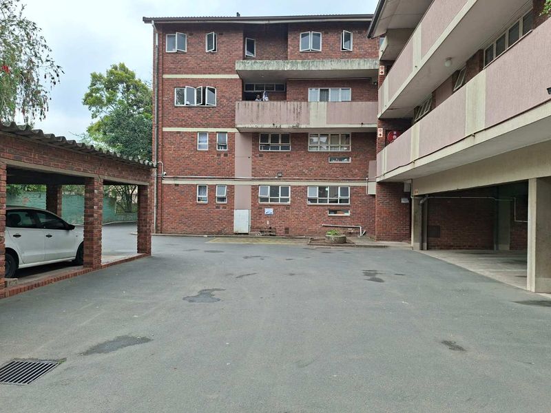 Exterior view of a multi-story brick apartment building featuring covered parking bays and an asphalt driveway area.