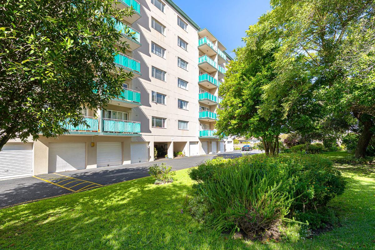 Exterior view of a multi-story apartment building featuring ground-level garage access, private balconies with green railings, and surrounding landscaped garden areas.