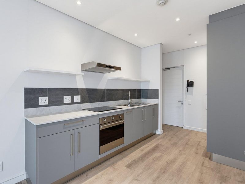 Modern, minimalist kitchen featuring grey cabinetry, stone countertops, integrated electric hob, oven, extractor fan, and sleek tiled backsplash in a clean, neutral space.