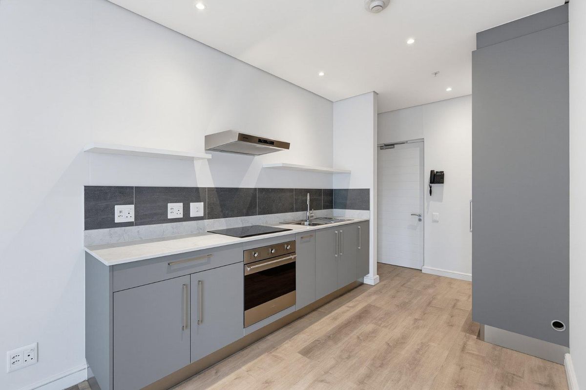 Modern, minimalist kitchen featuring grey cabinetry, stone countertops, integrated electric hob, oven, extractor fan, and sleek tiled backsplash in a clean, neutral space.